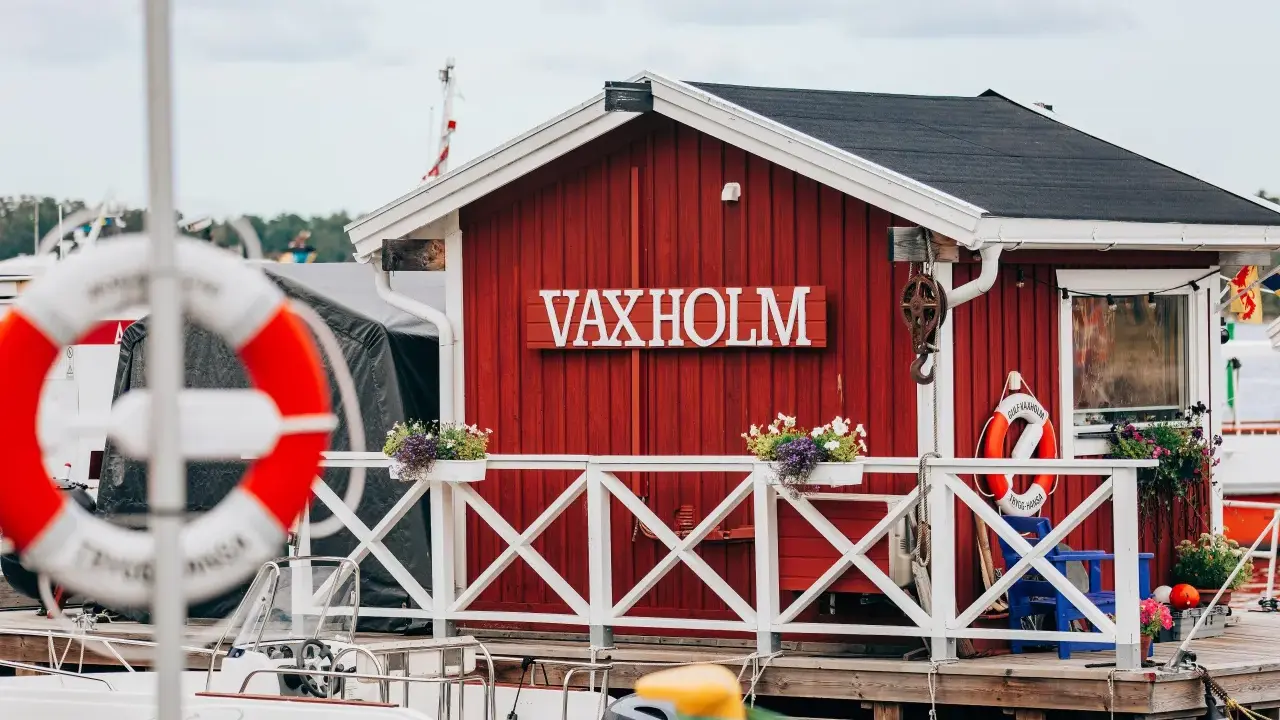 Rött hus med texten VAXHOLM vid vattnet. En livboj hänger på väggen. Vaxholms kastell skymtar i bakgrunden.