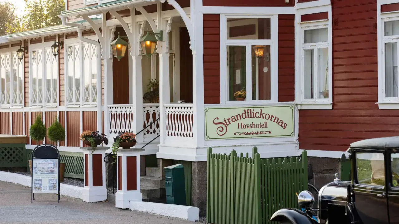 Strandflickornas Havshotell i Lysekil, en charmig röd byggnad med vita detaljer, välkomnar besökare. En klassisk bil står parkerad framför.