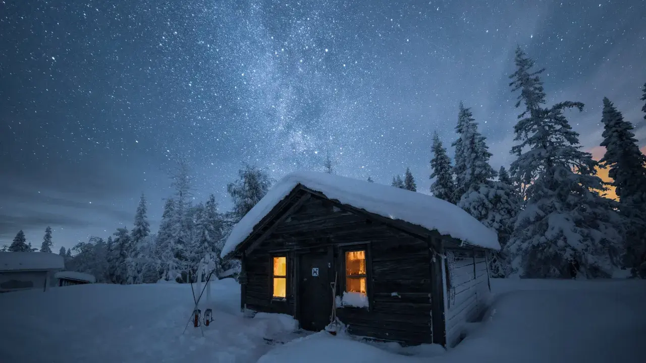 En snöig stuga under en stjärnklar himmel. Det är en perfekt kväll att fotografera vintergatan i Sverige.