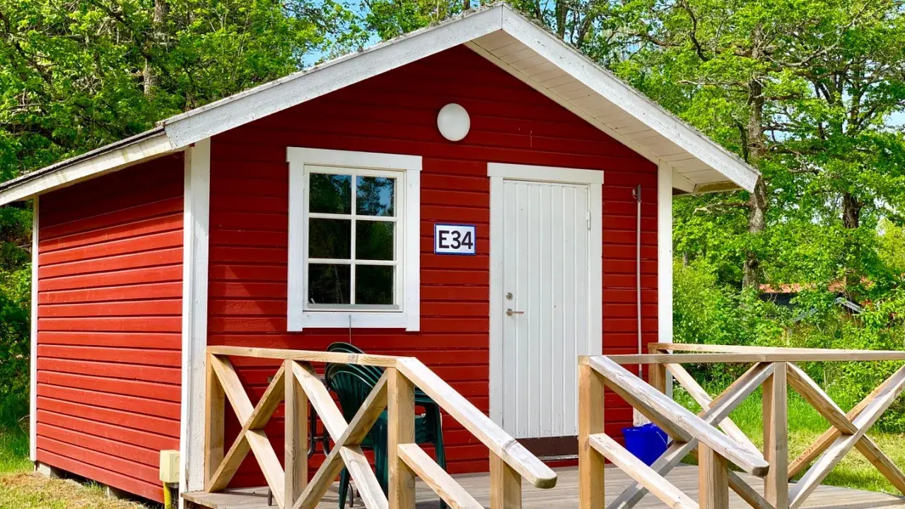 Rött litet hus med vit dörr och fönster, nummer E34. Trädäck med räcke. En grön stol står på däcket. Välkommen till Askeviks Camping!