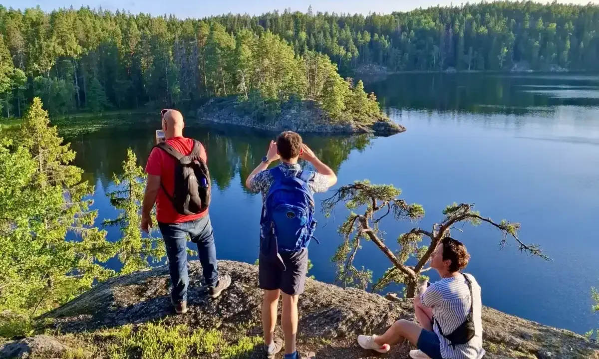 Vandring i Tyresta nationalpark. Tre personer njuter av utsikten över en stilla sjö och en tät skog. En man tar foto, en annan spanar ut, och en tredje vilar på en klippa.