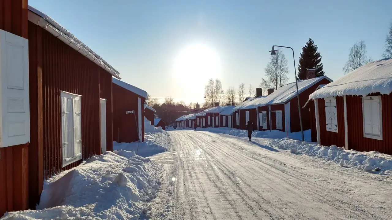 Vinterdag i kyrkbyn Gammelstad. Röda trähus med snö på taken kantar en snötäckt väg. Solen skiner starkt på en klarblå himmel.