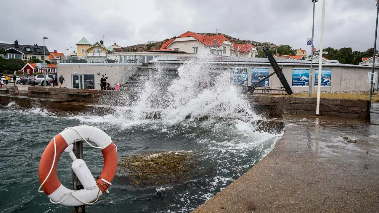 Vågor slår mot kajen i en kuststad, med hus i bakgrunden. En livboj syns i förgrunden. Det blåser med sekundmeter storm.