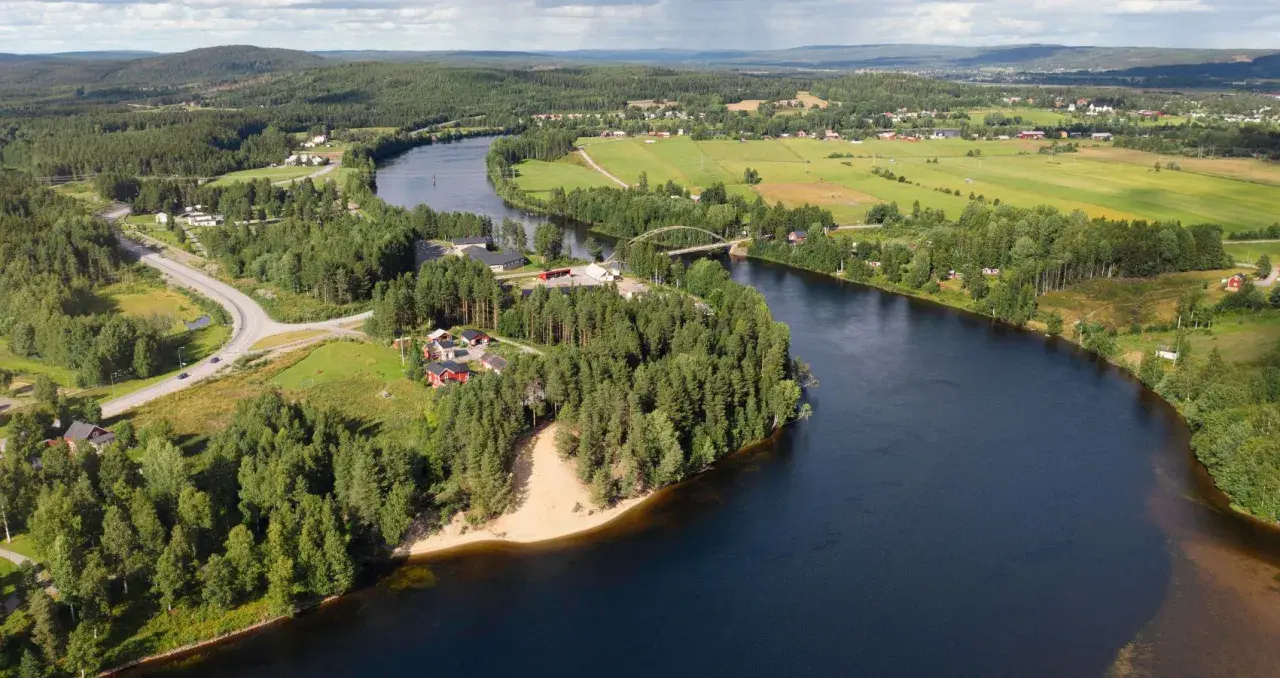 En vacker vy över en svensk landsbygd med en flod som slingrar sig genom gröna fält och skogar. En bro spänner över vattnet, och små röda hus syns bland träden. En idyllisk bild av sommar i Sverige.
