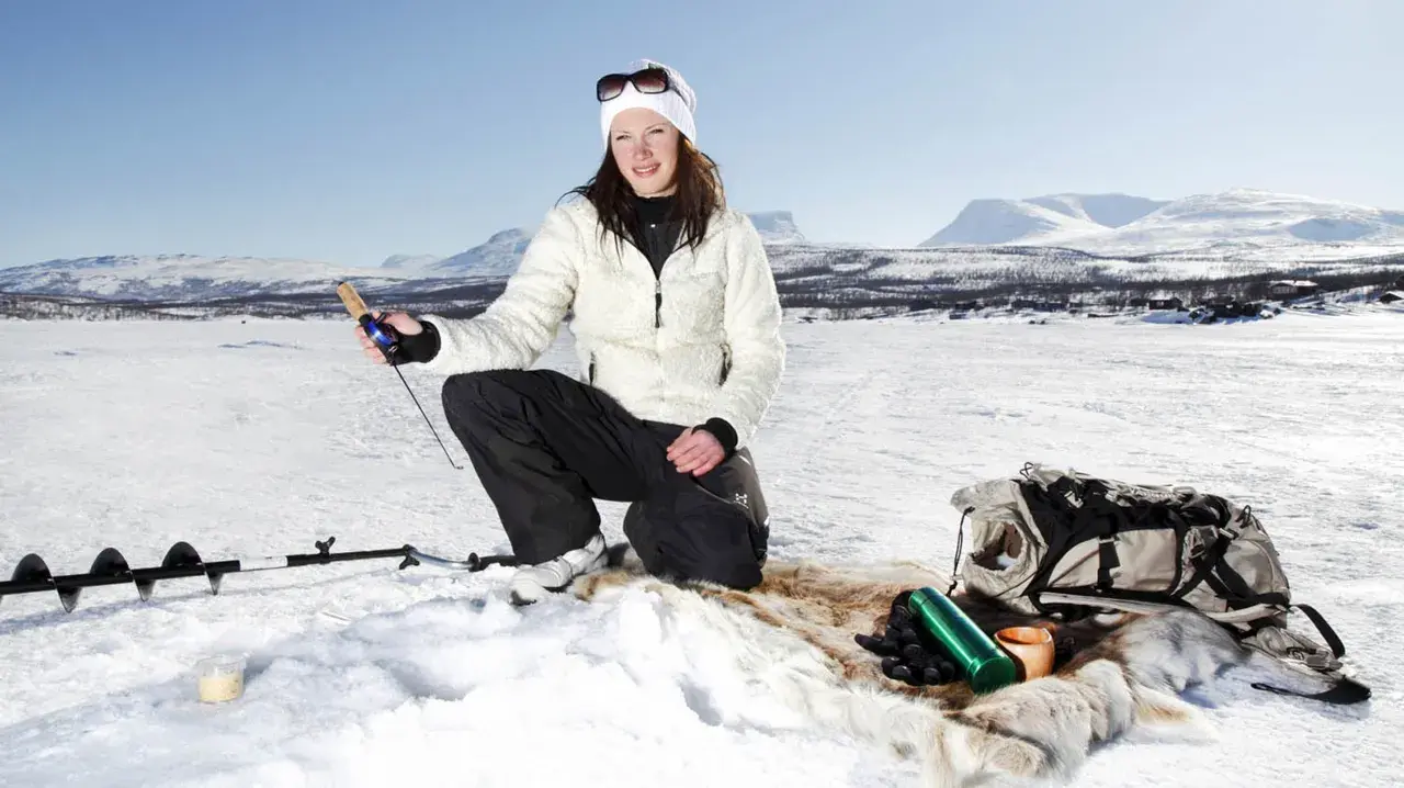 Kvinna ispirar isfiske i Kiruna. Hon har fiskeutrustning och en ryggsäck bredvid sig.
