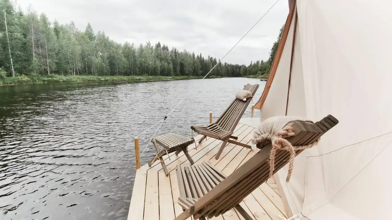 Två träfåtöljer och ett litet bord på en brygga vid en sjö, med skog i bakgrunden. En del av ett glampingtält syns. Upplev glamping Norrbotten.