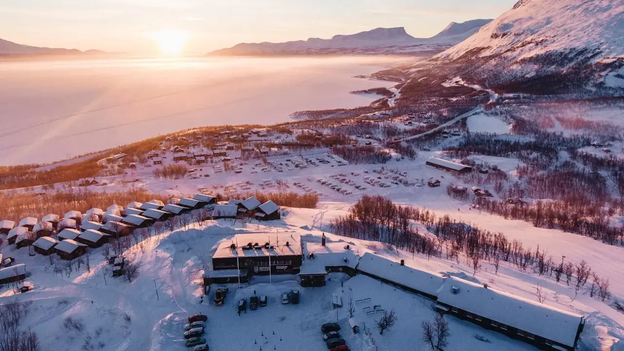 Vinterlandskap vid Kiruna med snötäckta fjäll, en frusen sjö och små stugor. Solen går ner och färgar himlen rosa.