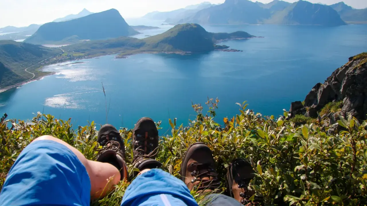 Fötter i vandringskängor vilar på en gräsbevuxen bergssluttning med en fantastisk utsikt över Lofoten. En perfekt dag för lofoten vandring.