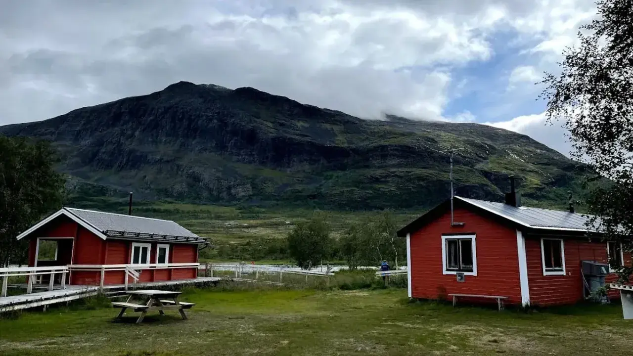 Två röda stugor i Abisko Nikkaluokta med en bergig, molntäckt bakgrund. En picknickbänk står på gräsmattan.