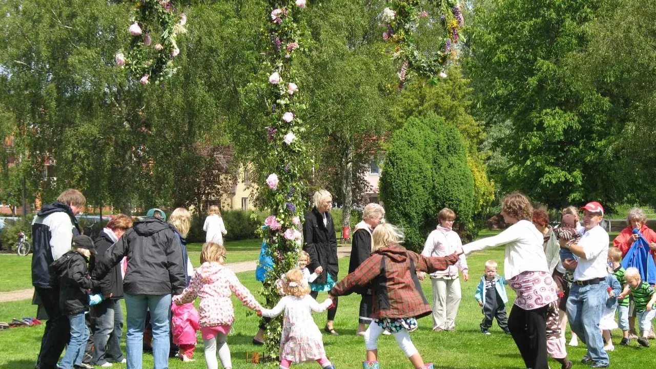 Barn och vuxna dansar runt en midsommarstång i en grönskande park.