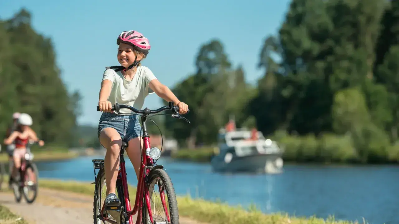 Glad flicka på cykel med hjälm njuter av en cykelsemester med barn längs en kanal. En båt syns i bakgrunden.