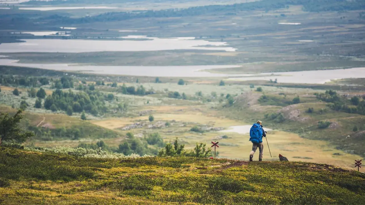 Vandrare med blå ryggsäck på Kungsleden, med vidsträckt landskap av sjöar och kullar.