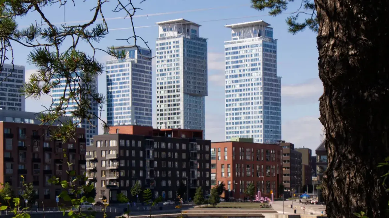 Höga, moderna skyskrapor reser sig bakom tegelbyggnader. En grönskande gren ramar in vyn, som om den undrar när sommaren startar i Sverige.