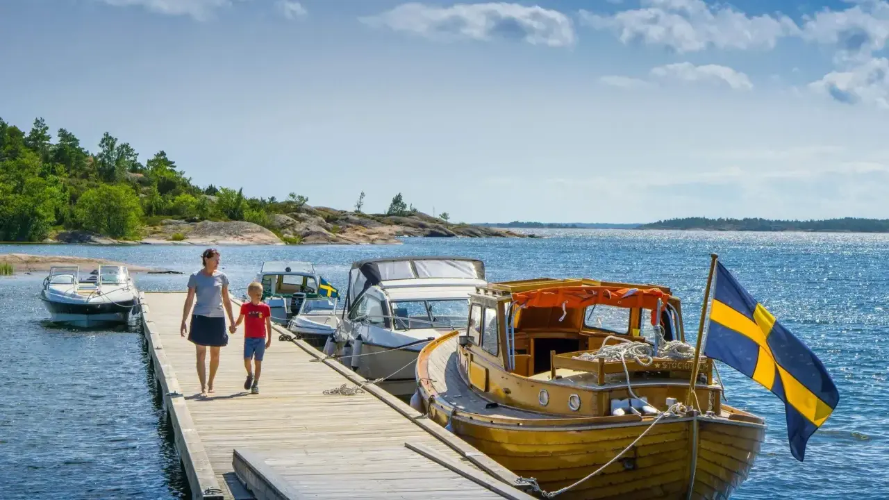 En kvinna och ett barn går på en brygga vid en sjö i Sverige. Båtar ligger förtöjda, en med svensk flagga. En perfekt familjesemester i Sverige.