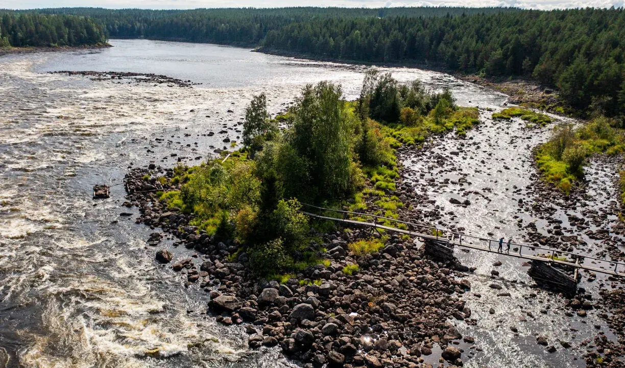 Vattenfall och en träbro över en stenig flod i Kalix, Sverige. Två personer går över bron.