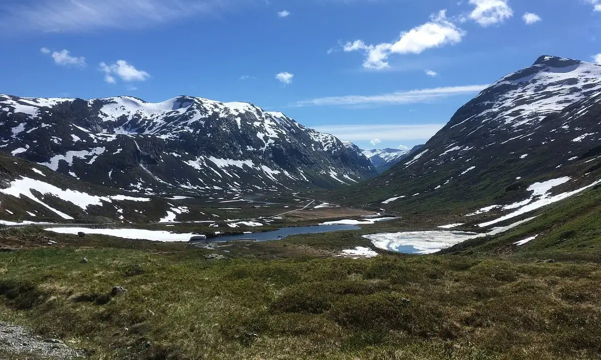 Vacker dalgång med snöklädda berg, en sjö och grönskande sluttningar. En väg slingrar sig fram, kanske en del av trollväggen.