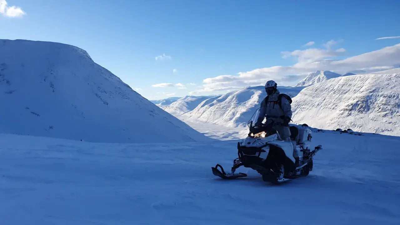 Snöskoterförare i Kebnekaise-området, med snöklädda berg och en klarblå himmel. En känsla av vidsträckthet och äventyr.