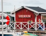 Rött hus med texten VAXHOLM vid vattnet. En livboj hänger på väggen. Vaxholms kastell skymtar i bakgrunden.