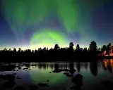 Norrsken dansar över en vinterlandskap vid Kiruna. En skogskant och en sjö med stenar speglar himlens magiska ljus.