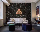 En mysig lounge med blommig tapet, en grå soffa, färgglada sittpuffar och en hylla full med böcker. En affisch för "Josef Frank - Against Design" hänger på väggen.