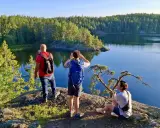 Vandring i Tyresta nationalpark. Tre personer njuter av utsikten över en stilla sjö och en tät skog. En man tar foto, en annan spanar ut, och en tredje vilar på en klippa.