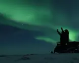 En person på snöskoter i Kiruna Lapland sträcker upp armarna mot norrskenet som lyser grönt på natthimlen.