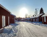 Vinterdag i kyrkbyn Gammelstad. Röda trähus med snö på taken kantar en snötäckt väg. Solen skiner starkt på en klarblå himmel.