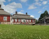 Malingsbo herrgård med sina röda trähus och stenfundament, omgivet av en grönskande äng med vita blommor under en blå himmel med moln.