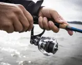 Händer som håller i ett fiskespö med en rulle, redo för fiska i Sverige. Vattnet glittrar i bakgrunden.