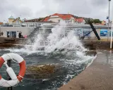 Vågor slår mot kajen i en kuststad, med hus i bakgrunden. En livboj syns i förgrunden. Det blåser med sekundmeter storm.
