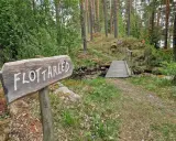 Träskylt med texten "Flottarleden Munkedal" pekar mot en liten träbro över en bäck i en skogsmiljö.