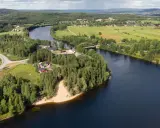En vacker vy över en svensk landsbygd med en flod som slingrar sig genom gröna fält och skogar. En bro spänner över vattnet, och små röda hus syns bland träden. En idyllisk bild av sommar i Sverige.
