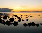 Solnedgång över havet vid en stenig strand på ar Gotland. Mörka stenar speglas i det lugna vattnet under en himmel i orange och gult.