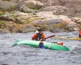 Två män i kajaker nära en klippig kust. En person i en grön kajak verkar ha problem. En diskussion om kanot vs kajak kan uppstå.