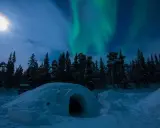 En glasigloo i Sverige under norrsken. En snöig skog och en fullmåne lyser upp natten.