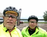 Två cyklister redo för Vätternrundan. Mannen har en keps med texten "Vätternrundan". Bakom dem syns en sjö och en flagga för Sollers IF Fotboll.