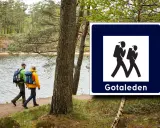 Två vandrare följer en stig vid en sjö, med en skylt för Gotaleden etapper.