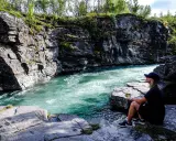 En kvinna sitter vid en forsande turkos flod i en klippig ravin. Vacker natur, kanske en vy över var ligger Abisko.