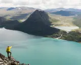 En person i gul jacka pekar ut över en turkos sjö och berglandskap under en Jotunheimen vandring.