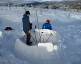 Två personer bygger en bivack i snön. De använder snöblock för att bygga väggarna och stavar för att markera utrymmet.
