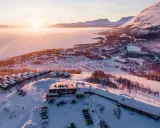 Vinterlandskap vid Kiruna med snötäckta fjäll, en frusen sjö och små stugor. Solen går ner och färgar himlen rosa.