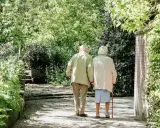 Ett äldre par går hand i hand ut på tur i en lummig park.