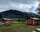 Två röda stugor i Abisko Nikkaluokta med en bergig, molntäckt bakgrund. En picknickbänk står på gräsmattan.