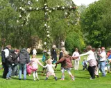 Barn och vuxna dansar runt en midsommarstång i en grönskande park.