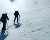 Två personer vandra på vintern uppför en snöklädd sluttning med skidutrustning och ryggsäckar.