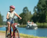 Glad flicka på cykel med hjälm njuter av en cykelsemester med barn längs en kanal. En båt syns i bakgrunden.