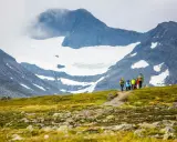 Vandringsgrupp på väg uppför en gräsbeklädd sluttning med stenar, mot en glaciärklädd bergstopp. En spännande utmaning att bestiga Sylarna.