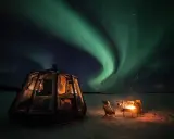 En glaskupol för camping i Norrland lyser upp natten under ett dansande norrsken. En eld sprakar vid sidan om.
