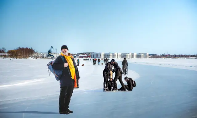 Glad man med ryggsäck njuter av en solig dag på isen i Luleå. Folk åker skridskor och promenerar i bakgrunden.
