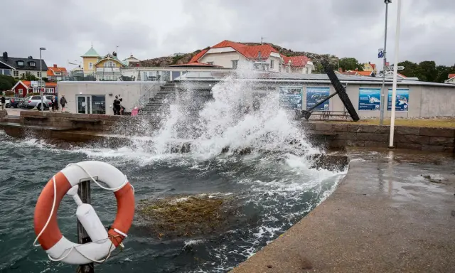 Vågor slår mot kajen i en kuststad, med hus i bakgrunden. En livboj syns i förgrunden. Det blåser med sekundmeter storm.